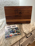 May include: A rectangular wooden cutting board with a dark brown finish, featuring a light wood accent stripe. The board is engraved with "Matt's Meat Shack" and a logo. A cookbook titled "Good Food" is also visible.